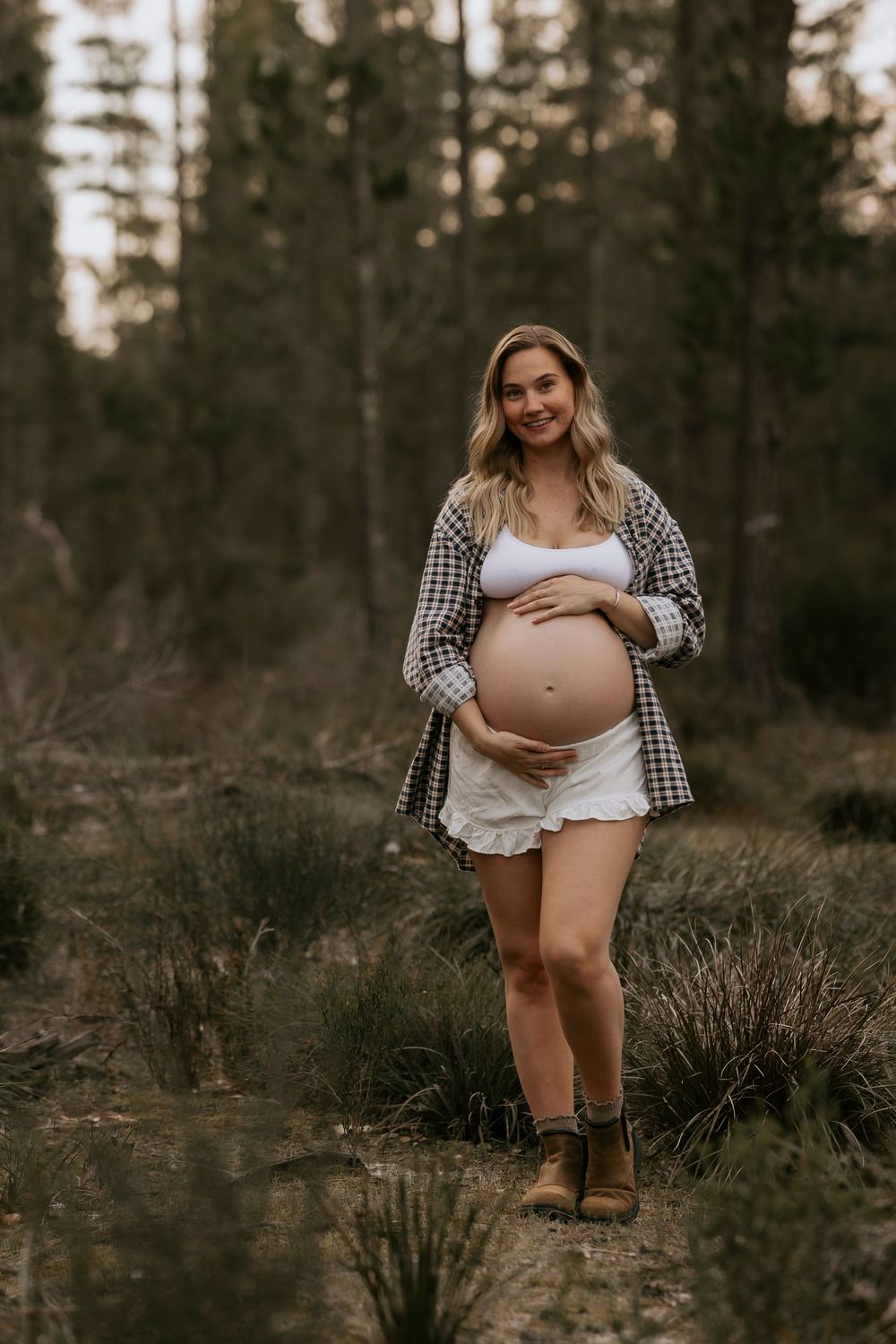 Smiling pregnant woman in plaid shirt and white shorts cradling baby bump in a forest setting.