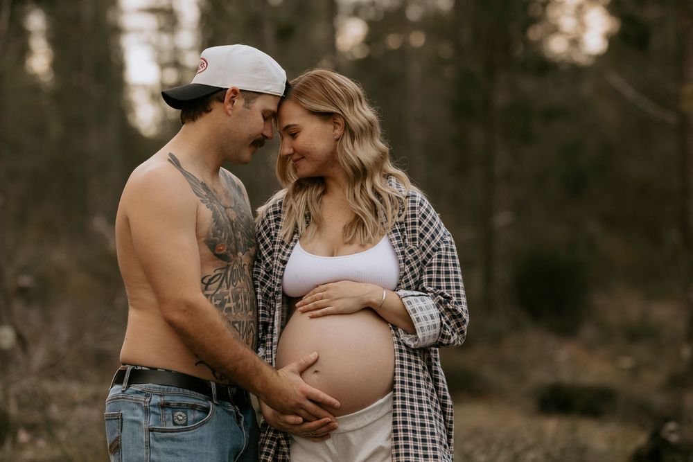 Shirtless tattooed man and pregnant woman embrace outdoors in forest setting for maternity photo shoot.