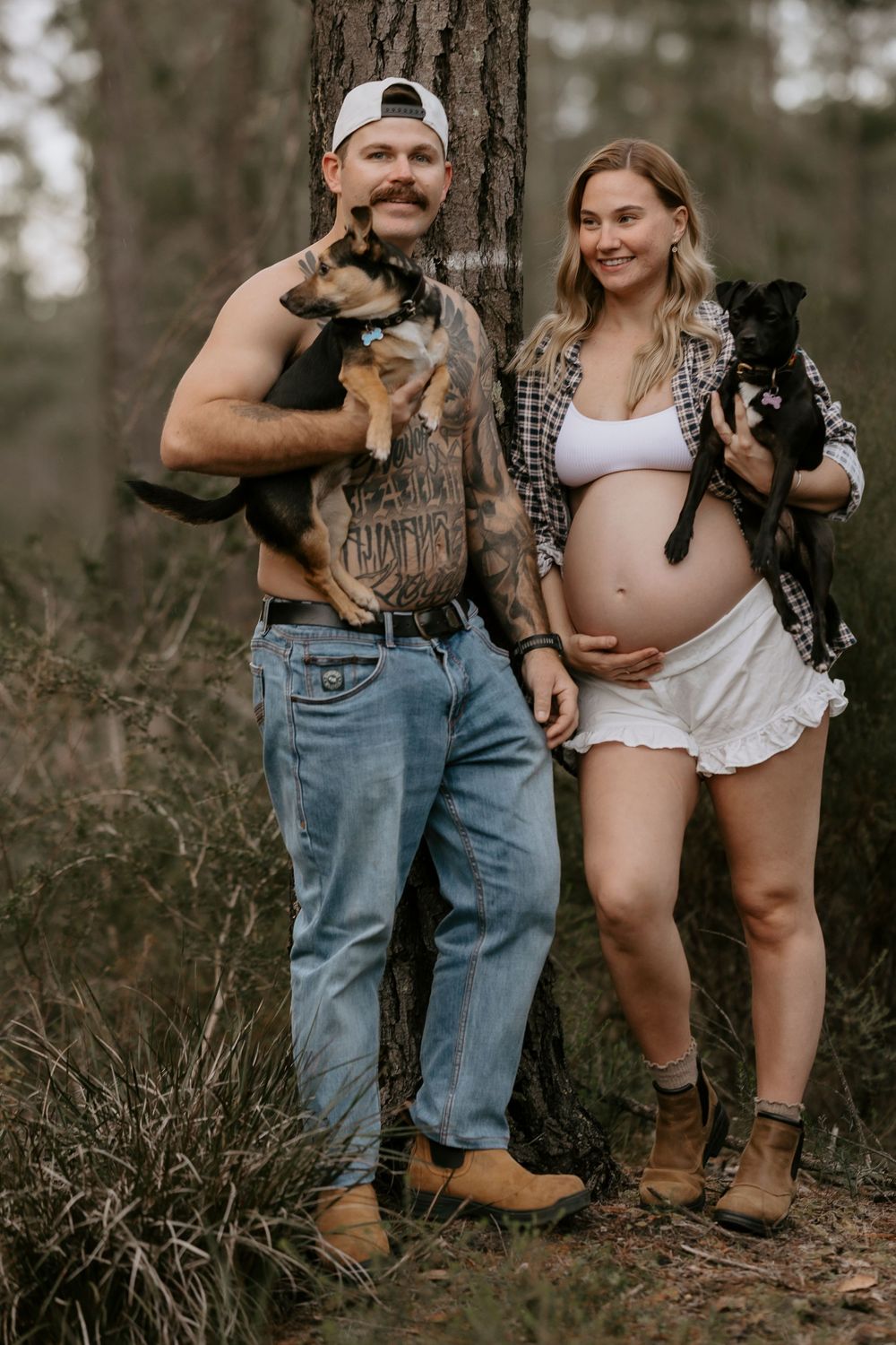 Smiling couple leaning against tree in forest, each holding a puppy, pregnant woman in white skirt.