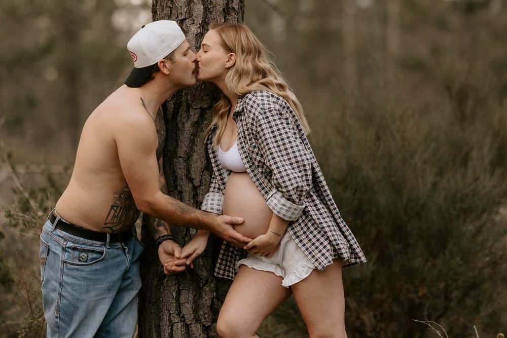 Couple kissing against a tree in a forest, woman in open plaid shirt showing pregnant belly outdoors.