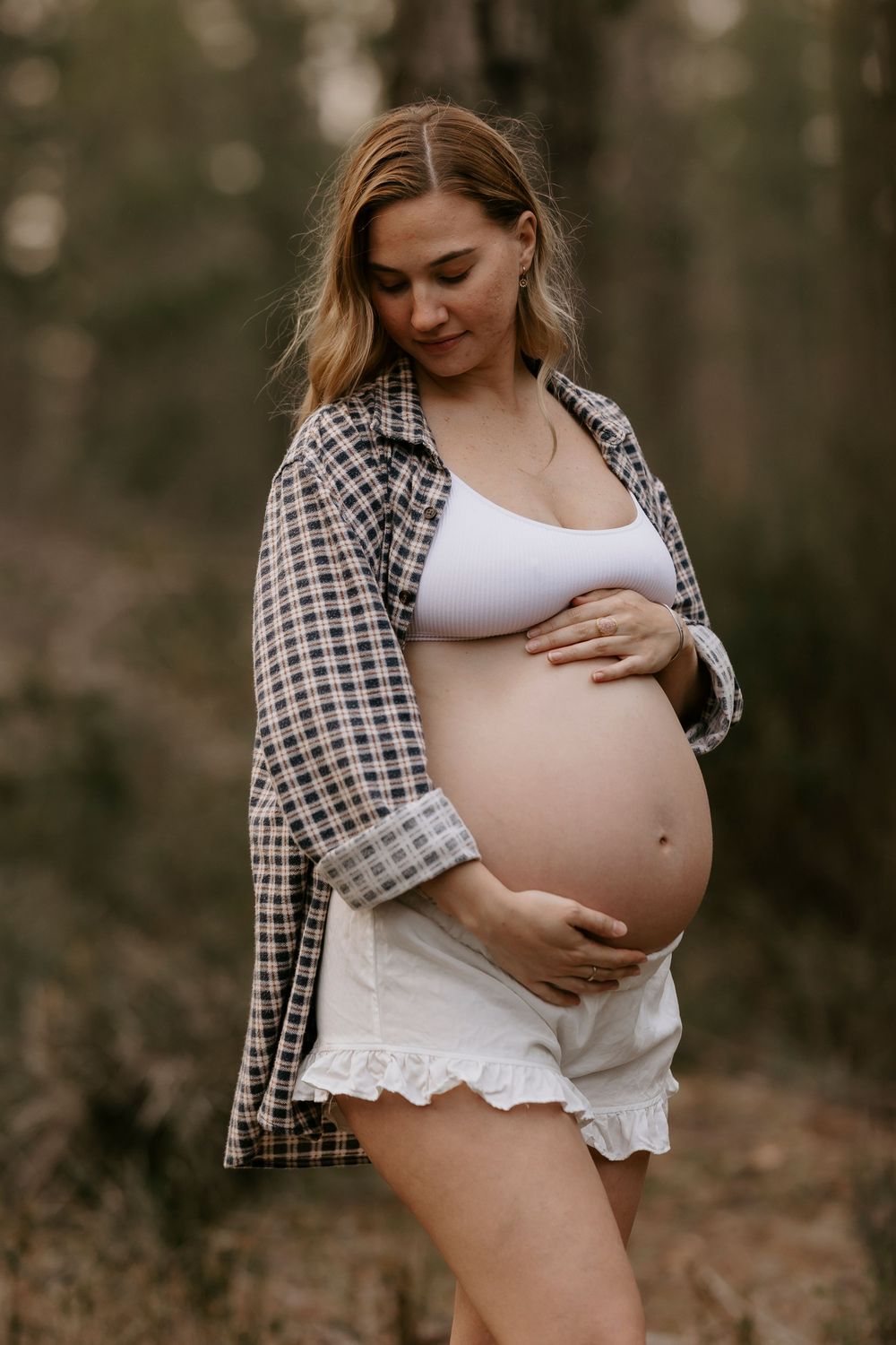 Pregnant woman in plaid shirt and white shorts tenderly holding her baby bump outdoors in woods.