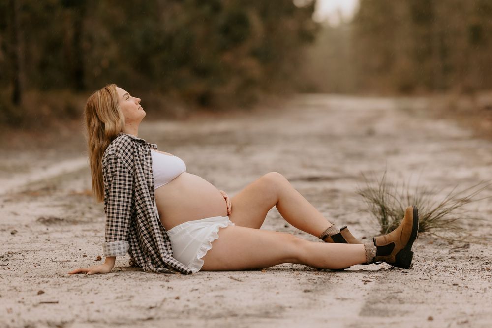 Pregnant woman in white outfit and plaid shirt sitting on a dirt road surrounded by trees during a maternity photoshoot.