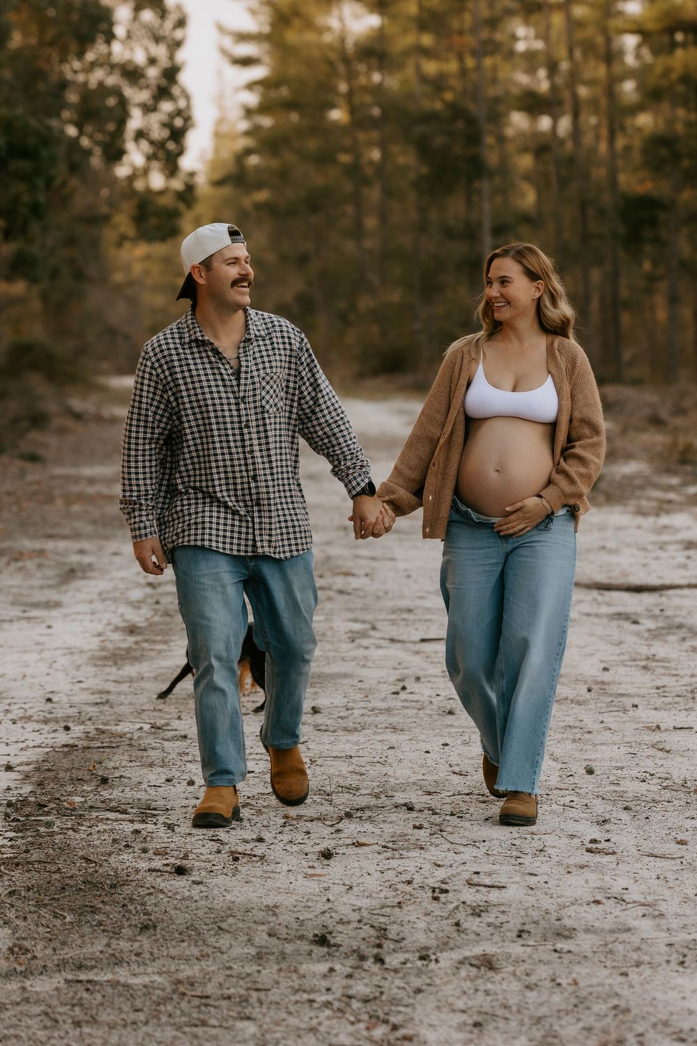 Couple holding hands walking on a dirt path through woods, pregnant woman in white crop top and jeans.