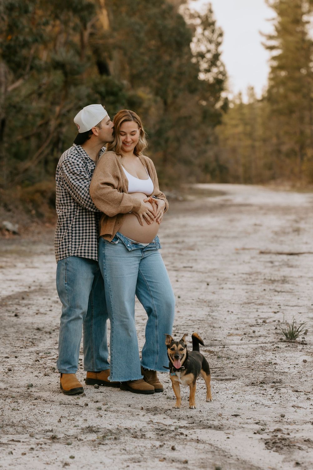 Couple sharing a tender moment on a sandy forest path, with the woman showing a baby bump and a small dog at their feet.