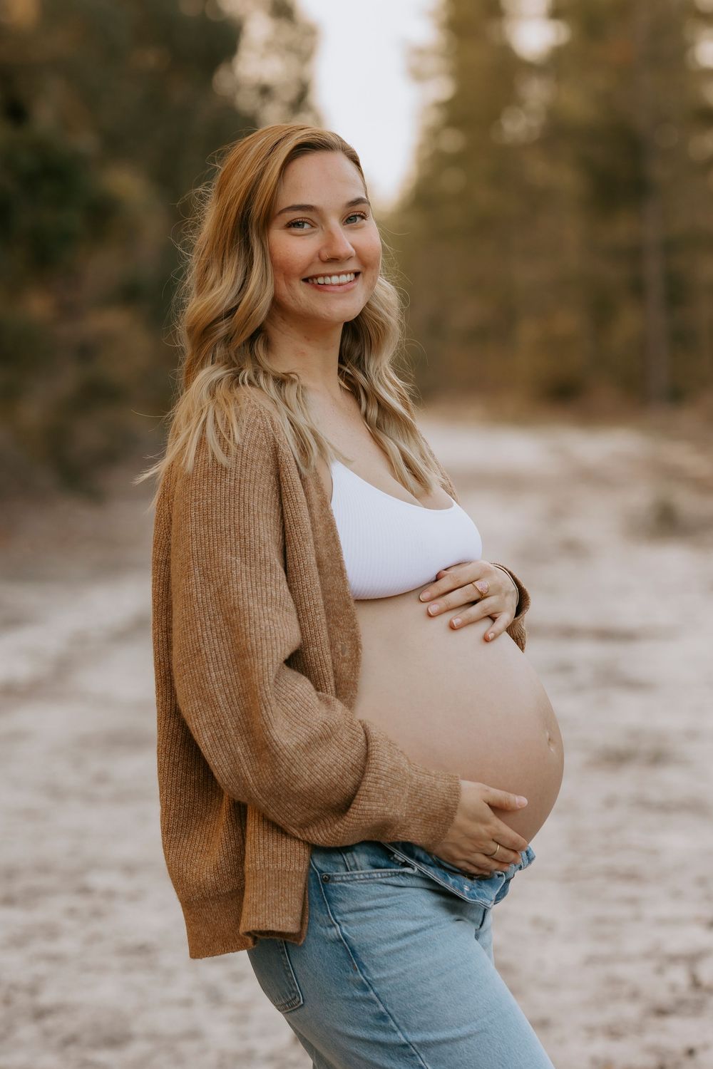 Smiling pregnant woman in brown cardigan and jeans cradling baby bump outdoors.