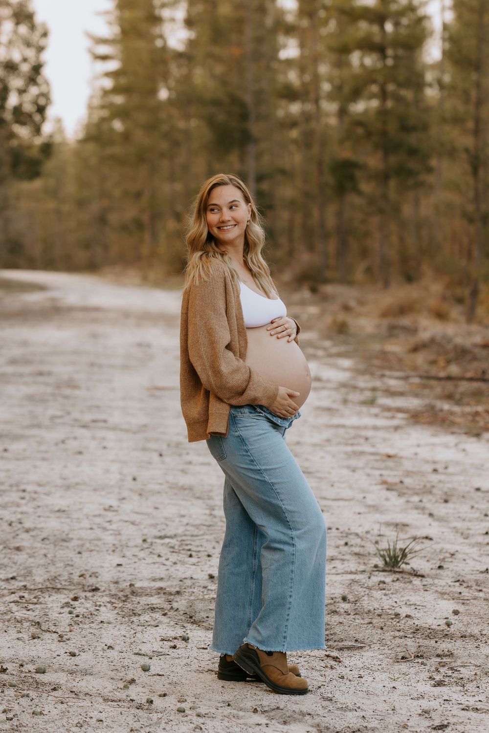 Smiling pregnant woman in jeans and brown cardigan cradling baby bump on a sandy forest path.