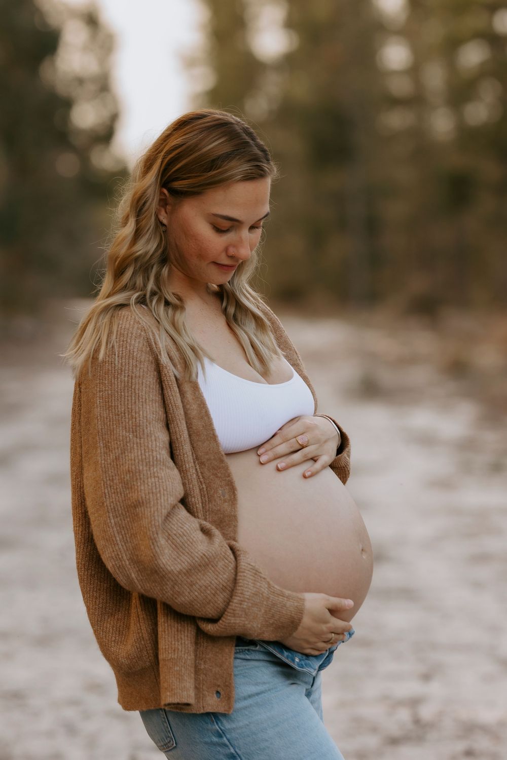 Pregnant woman in brown cardigan and jeans cradling baby bump outdoors on a sandy path surrounded by trees.