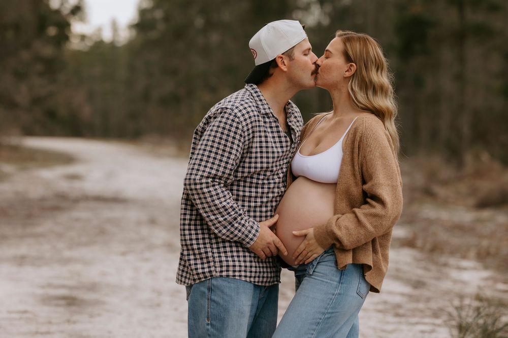 Couple sharing a kiss during a maternity photoshoot on a dirt road, with man cradling pregnant woman's belly.