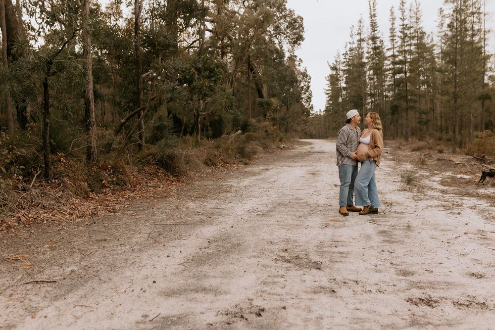 Couple embracing on a sandy forest path surrounded by tall pine trees during an outdoor maternity photo session.