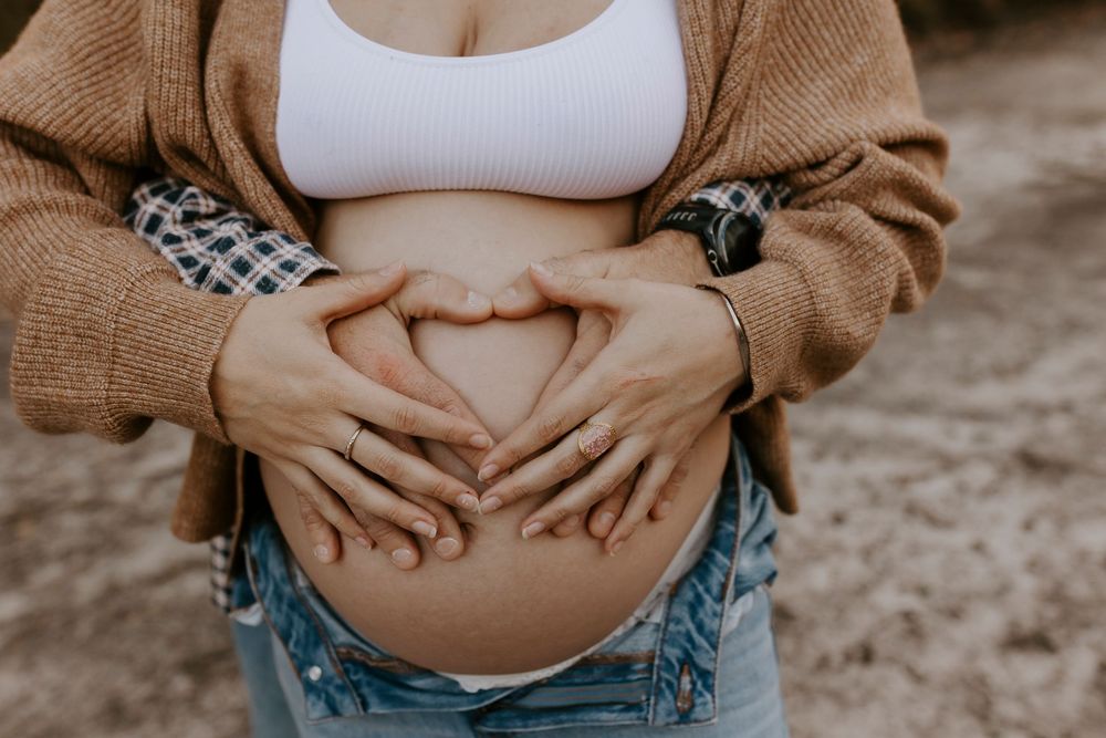Pregnant woman in white crop top and jeans forming heart shape on baby bump with hands outdoors.