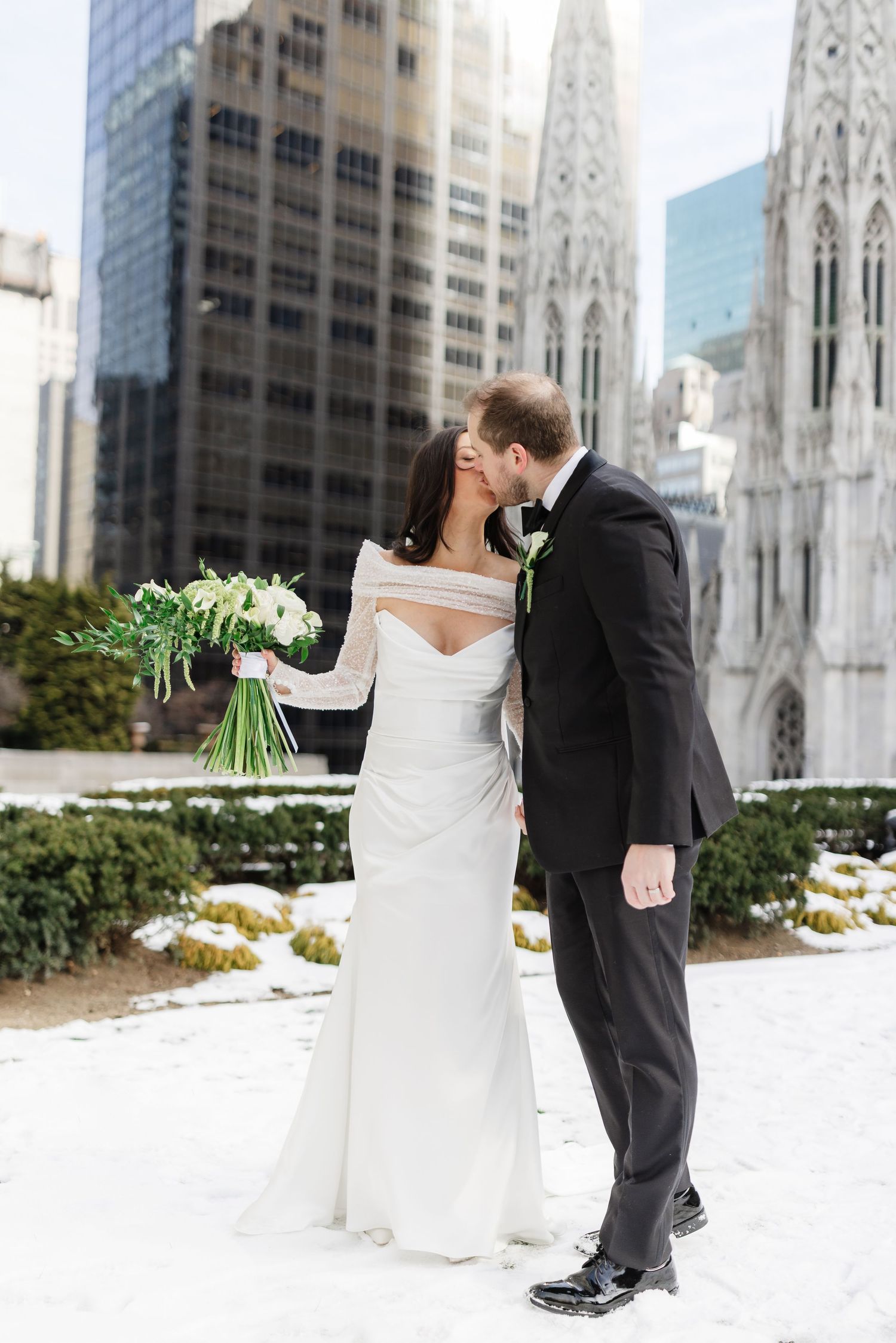 Bride and groom kissing in snowy NYC with St. Patrick's Cathedral and skyscrapers backdrop, bride holding white bouquet.