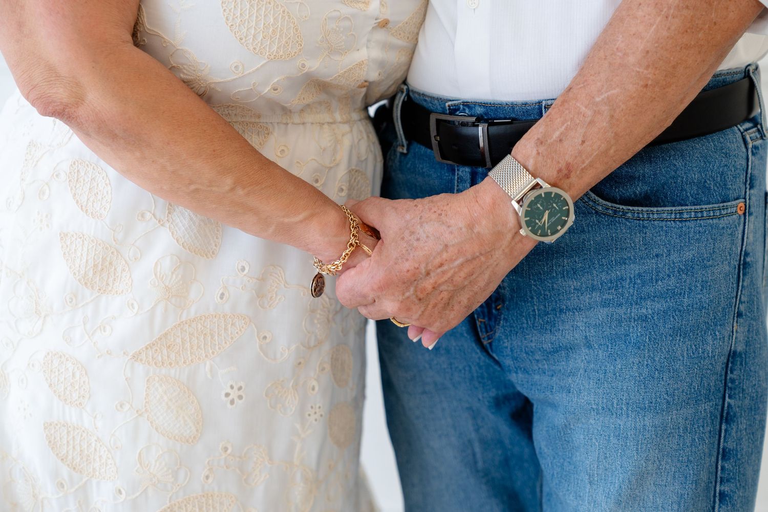 Elderly couple holding hands, woman in white lace dress with gold bracelet, man in jeans with green-faced watch.
