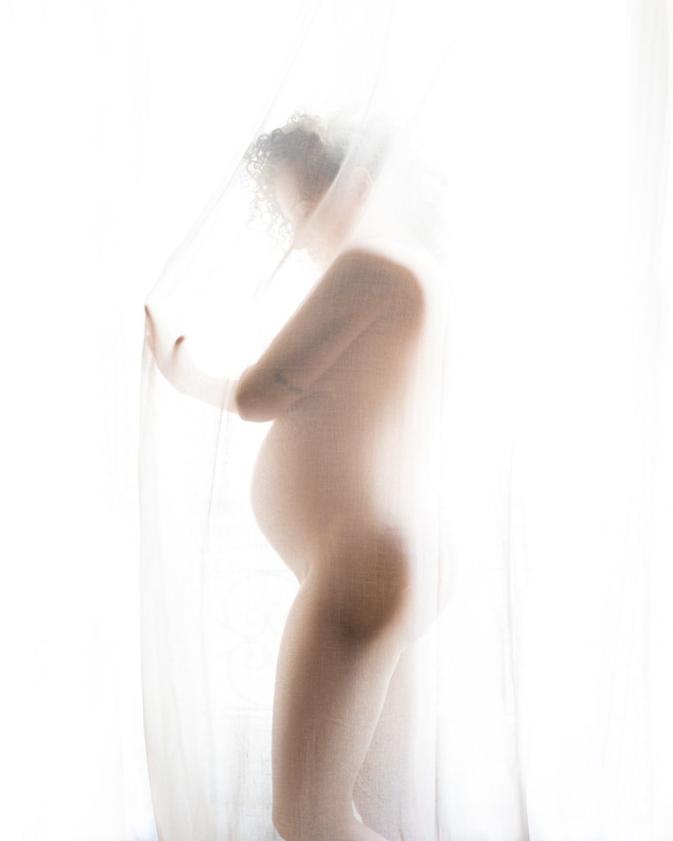 Silhouette of a pregnant woman draped in sheer white fabric, glowing in soft backlight for an ethereal maternity portrait.