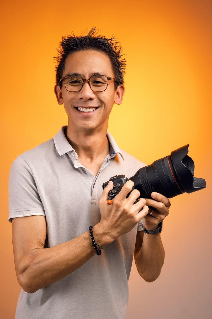 Smiling Asian man in glasses holding a DSLR camera against an orange background.