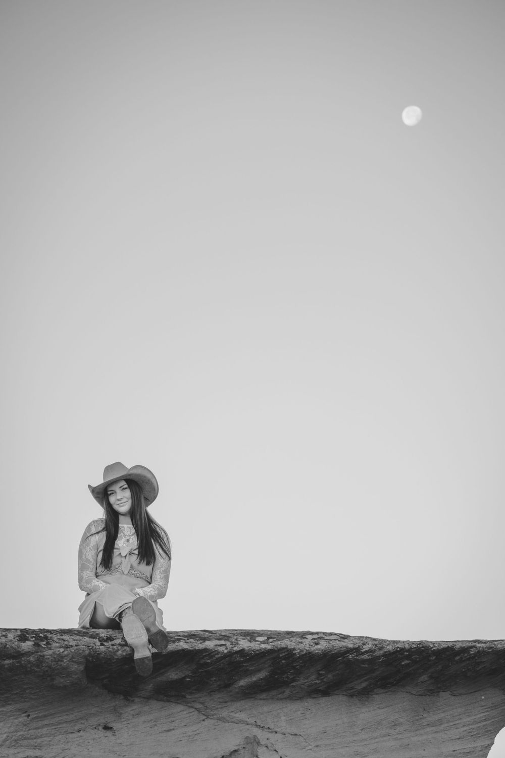 Black and white photo of a smiling woman in a cowboy hat sitting on a rock with the moon visible in the sky.