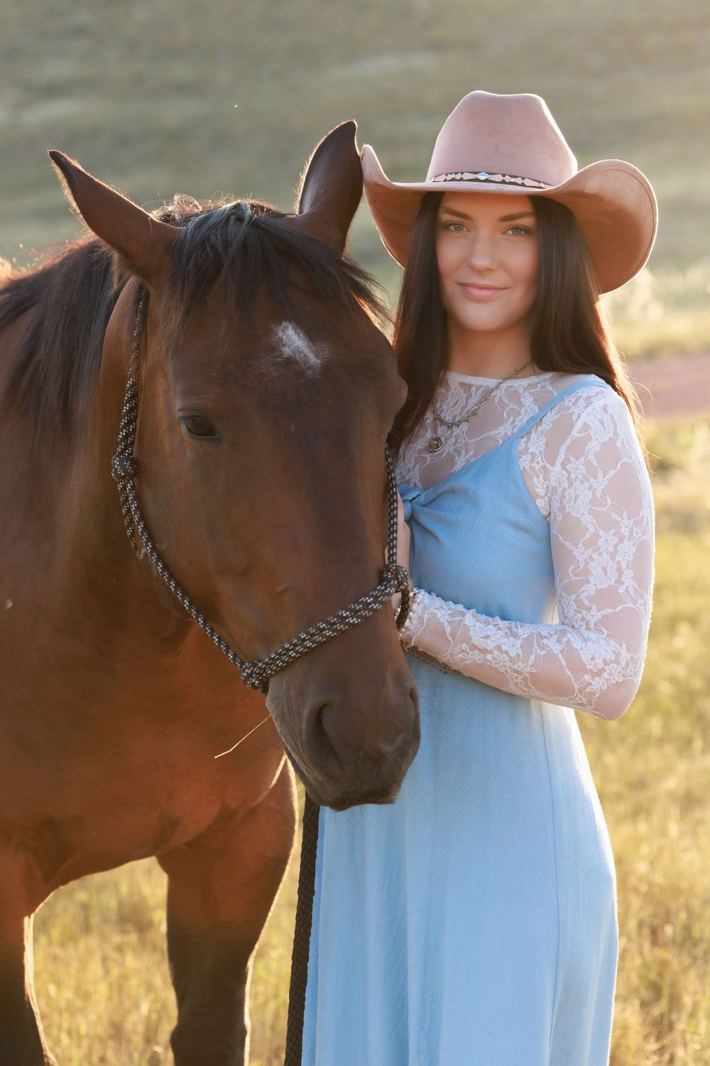 A young woman in a blue lace dress and pink cowboy hat stands beside a brown horse in a sunlit field.