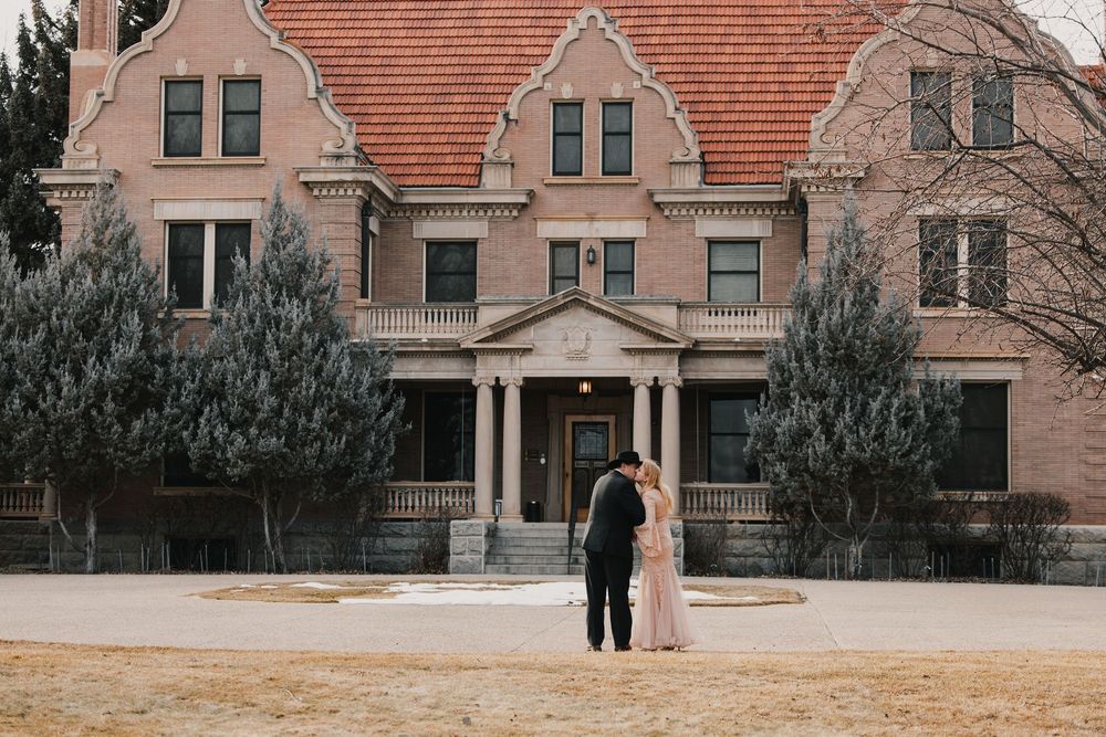 Couple embracing in front of a grand historic brick mansion with ornate architecture and evergreen trees.