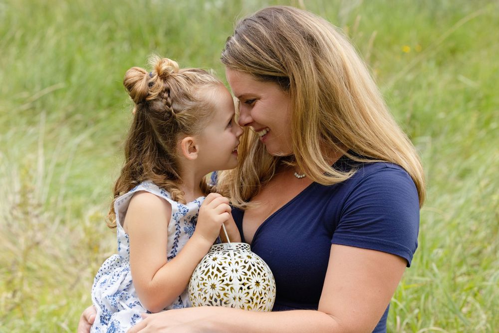 A mother smiles warmly at her young daughter outdoors in a green meadow, sharing a tender moment together.