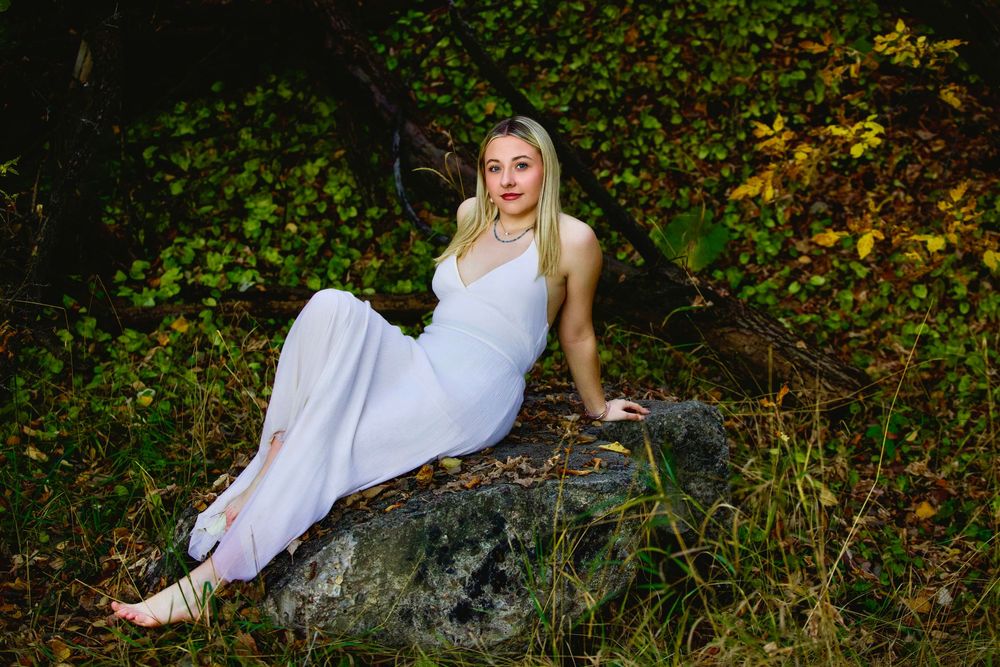 A blonde woman in a white dress sits gracefully on a rock surrounded by lush green forest foliage.