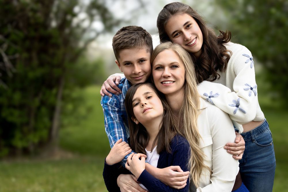 Four smiling young people pose together outdoors in a park, embracing warmly with green trees in the background.
