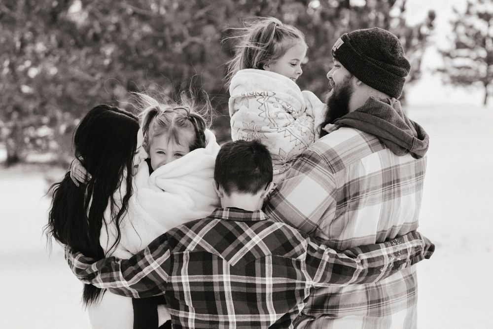Black and white photo of a family hugging outdoors in winter, parents holding young children in a snowy setting.