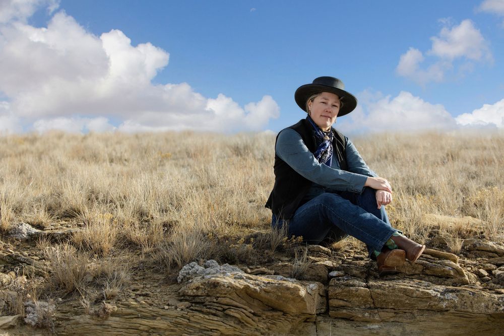 Person in cowboy hat sitting on rocks in a vast golden grassland under a blue sky with white clouds.