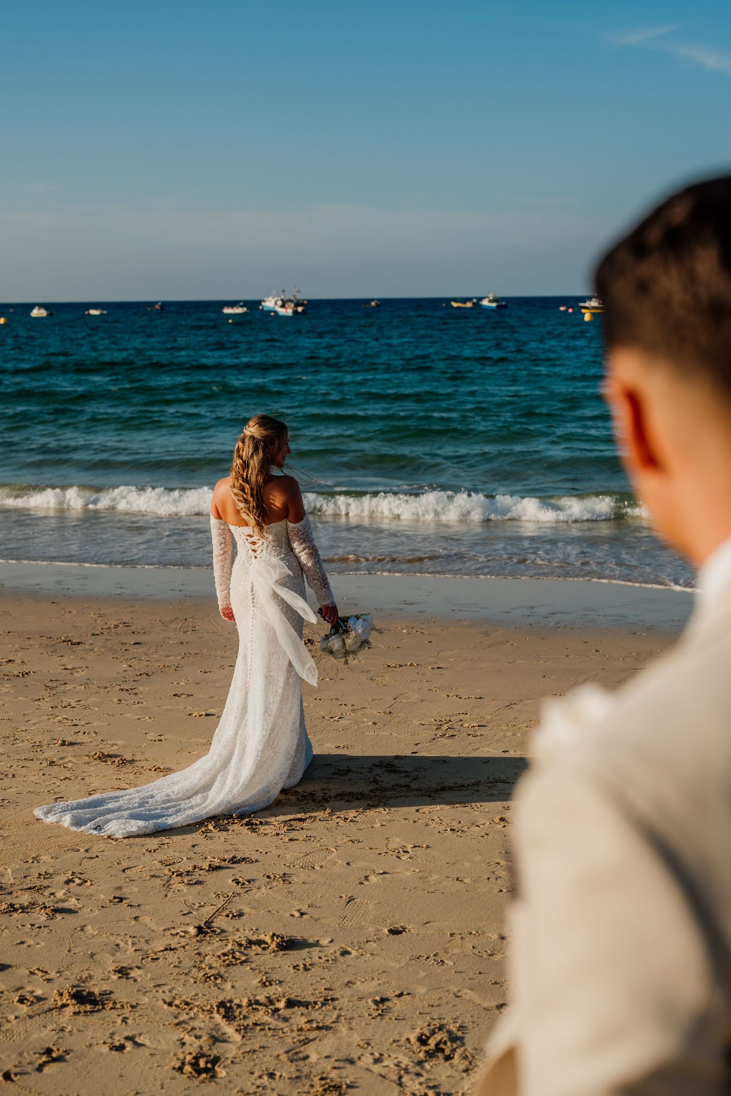 Bride in white lace gown holding bouquet stands at ocean's edge on sandy beach while groom watches in foreground.