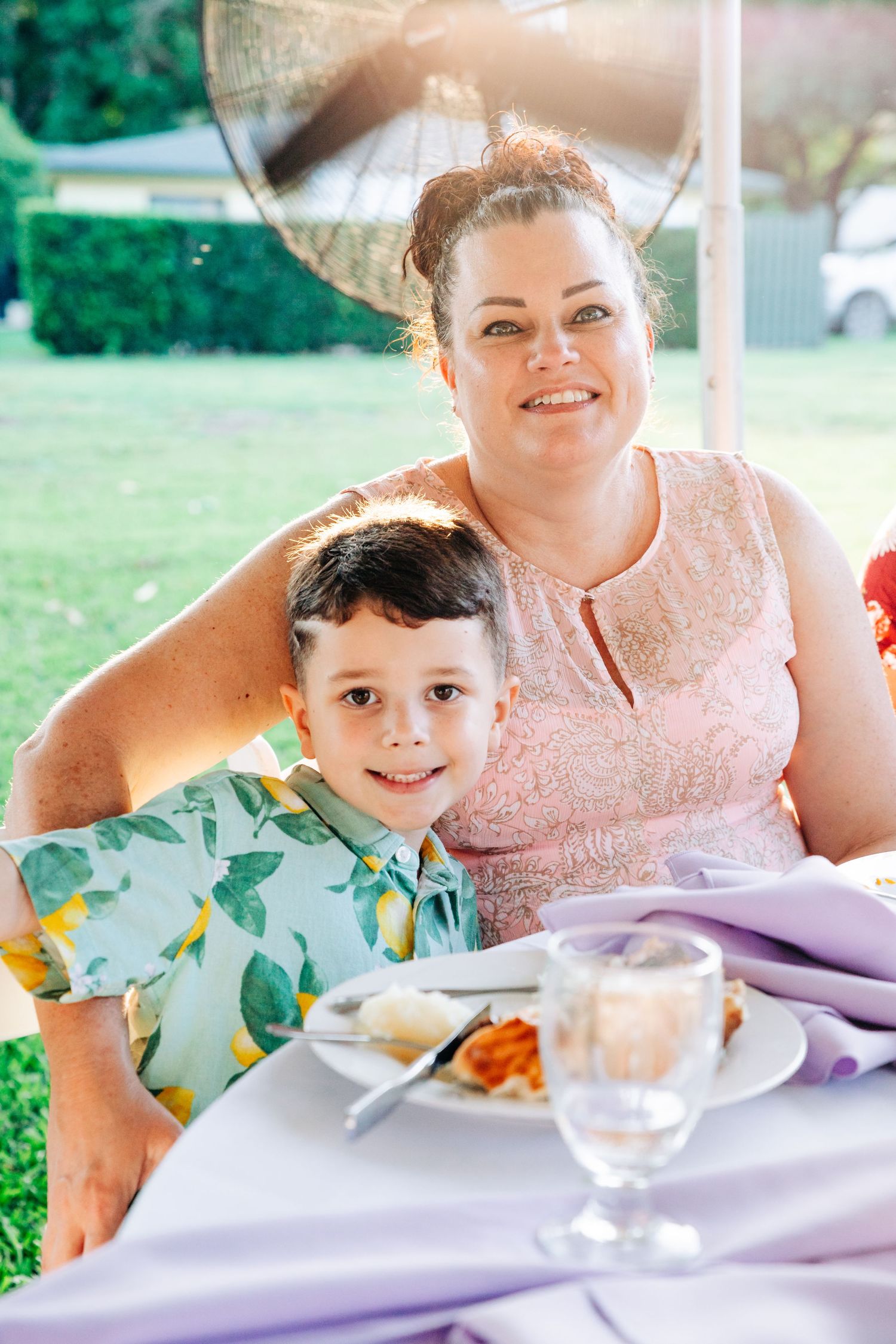 A woman and young boy smile together at an outdoor dining table set with food and drinks on a sunny day.