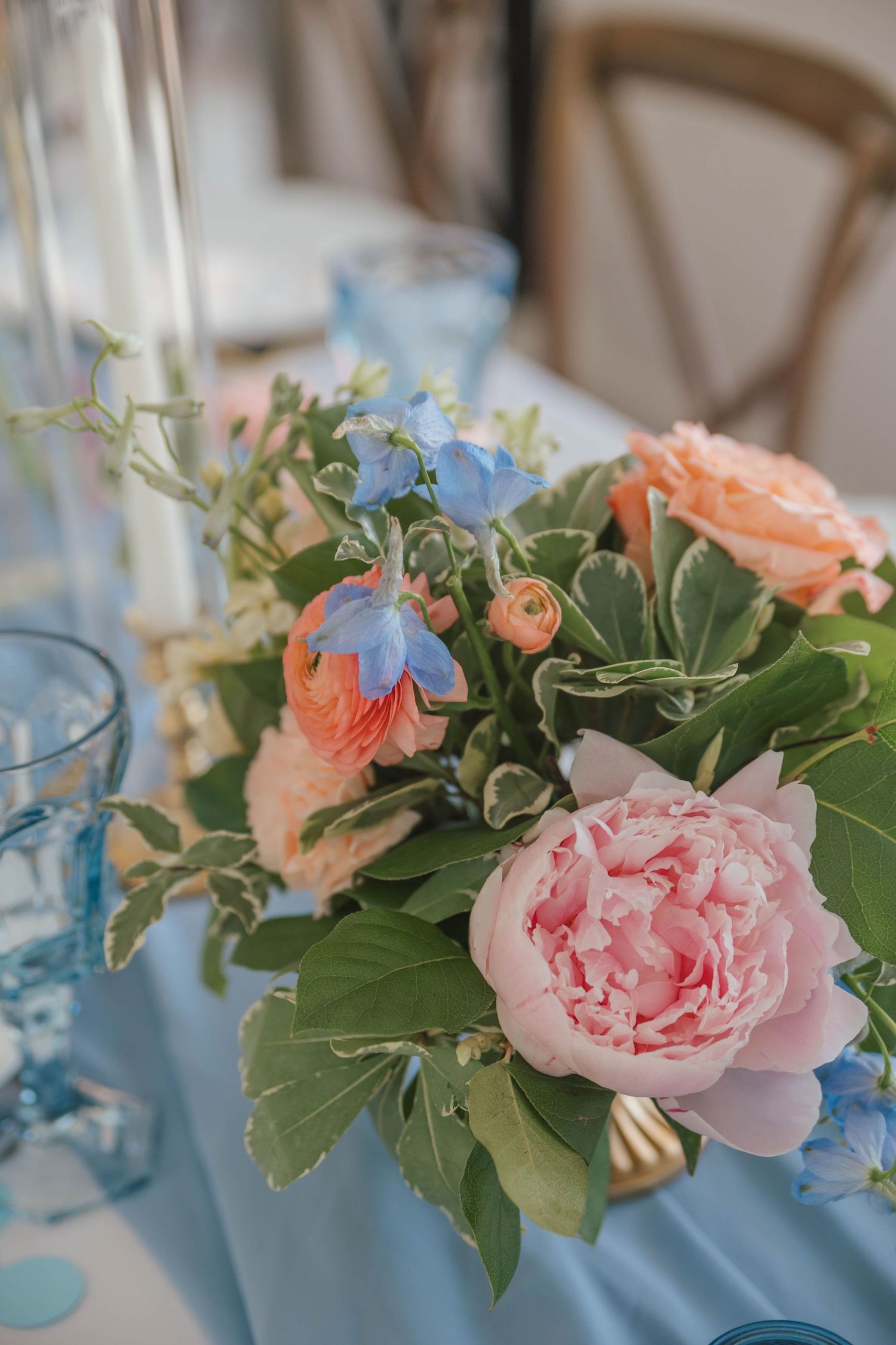Elegant floral centerpiece with pink peonies, peach roses, and blue flowers on a light blue tablecloth.