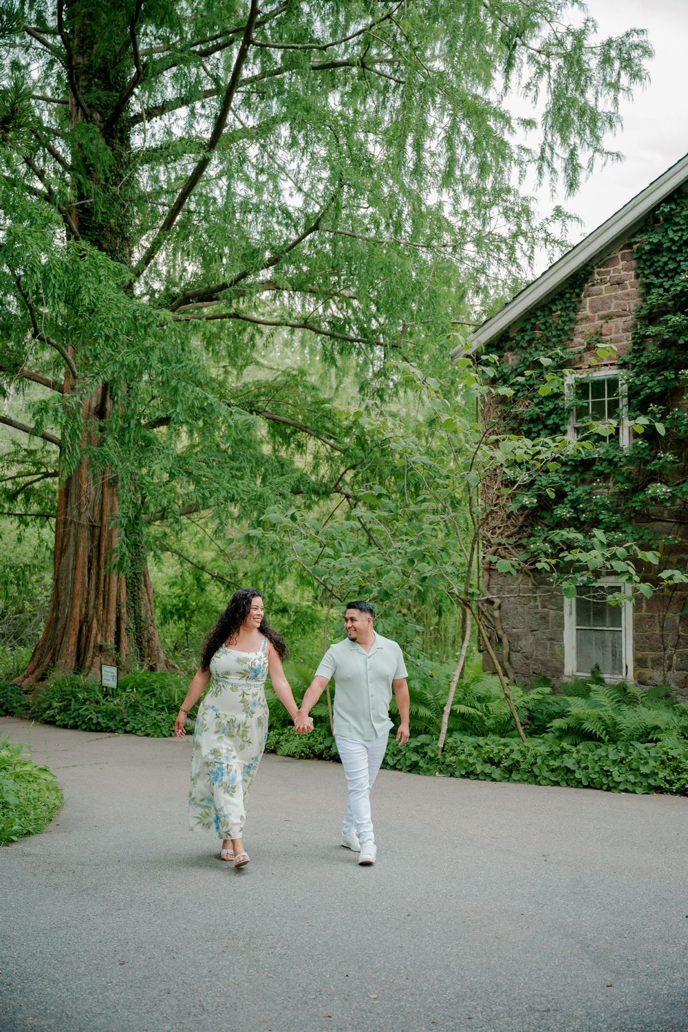 Couple holding hands walking along a garden path surrounded by lush greenery and an ivy-covered stone building.