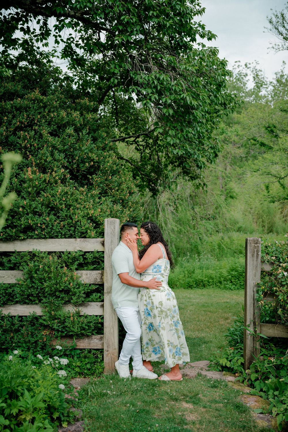Romantic portrait of an engaged couple leaning against a rustic wooden fence surrounded by greenery at Willowwood Arboretum, NJ.