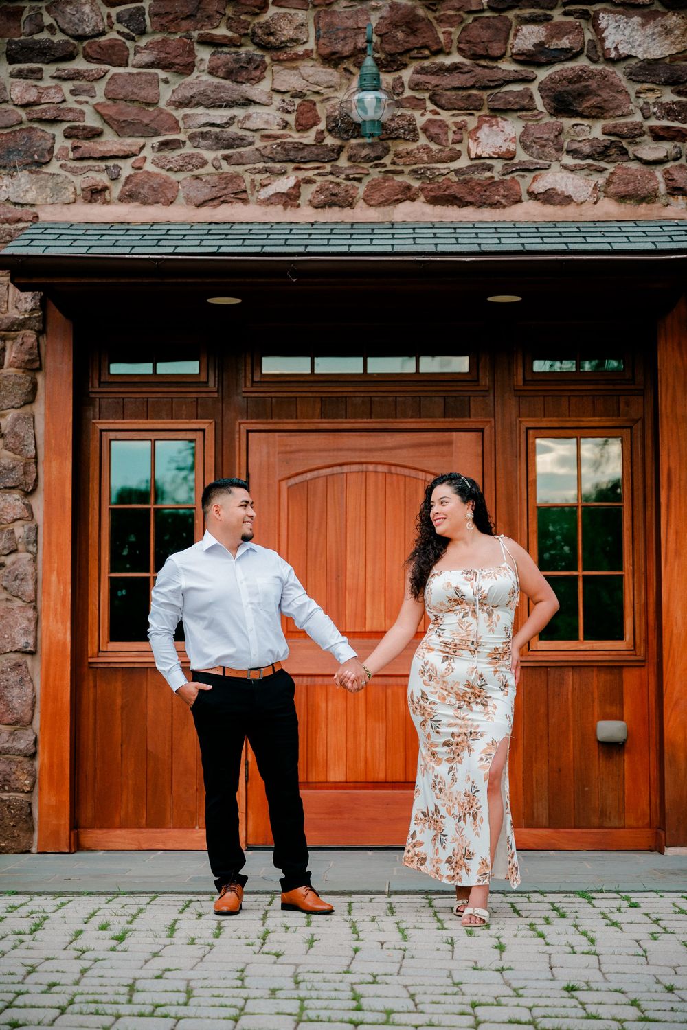 Couple holding hands in front of elegant wooden doors and stone wall, man in white shirt, woman in floral dress.