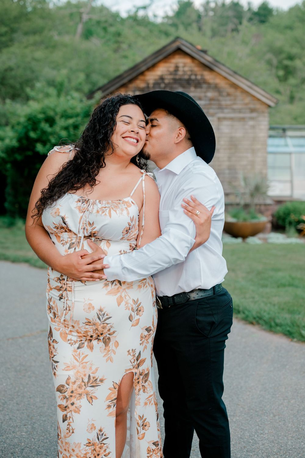 Couple embracing outdoors, woman in floral dress, man in black cowboy hat kissing her cheek near rustic barn.