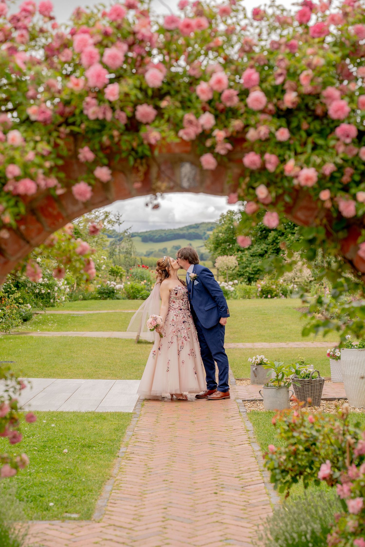 Couple kissing under a pink rose arch in a lush garden, bride in floral gown and groom in navy suit.