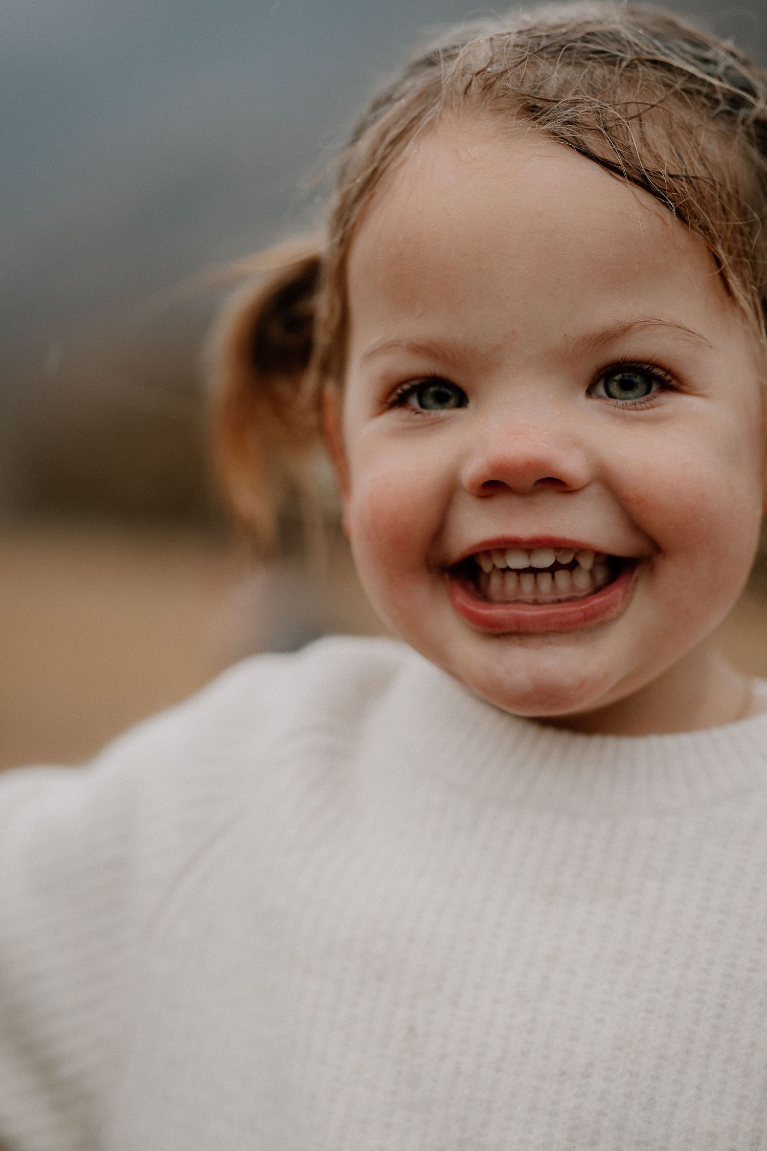 A joyful toddler girl with pigtails smiles broadly, wearing a white knit sweater outdoors.