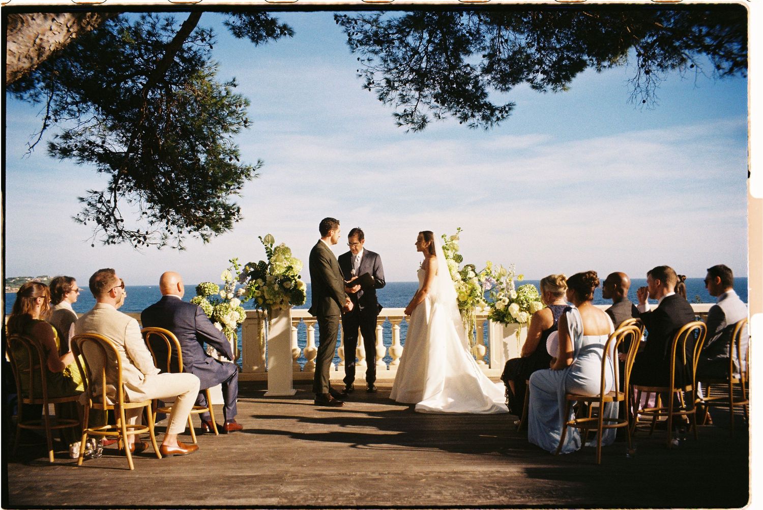 Panoramic view of an outdoor wedding ceremony at sunset with guests seated under trees.