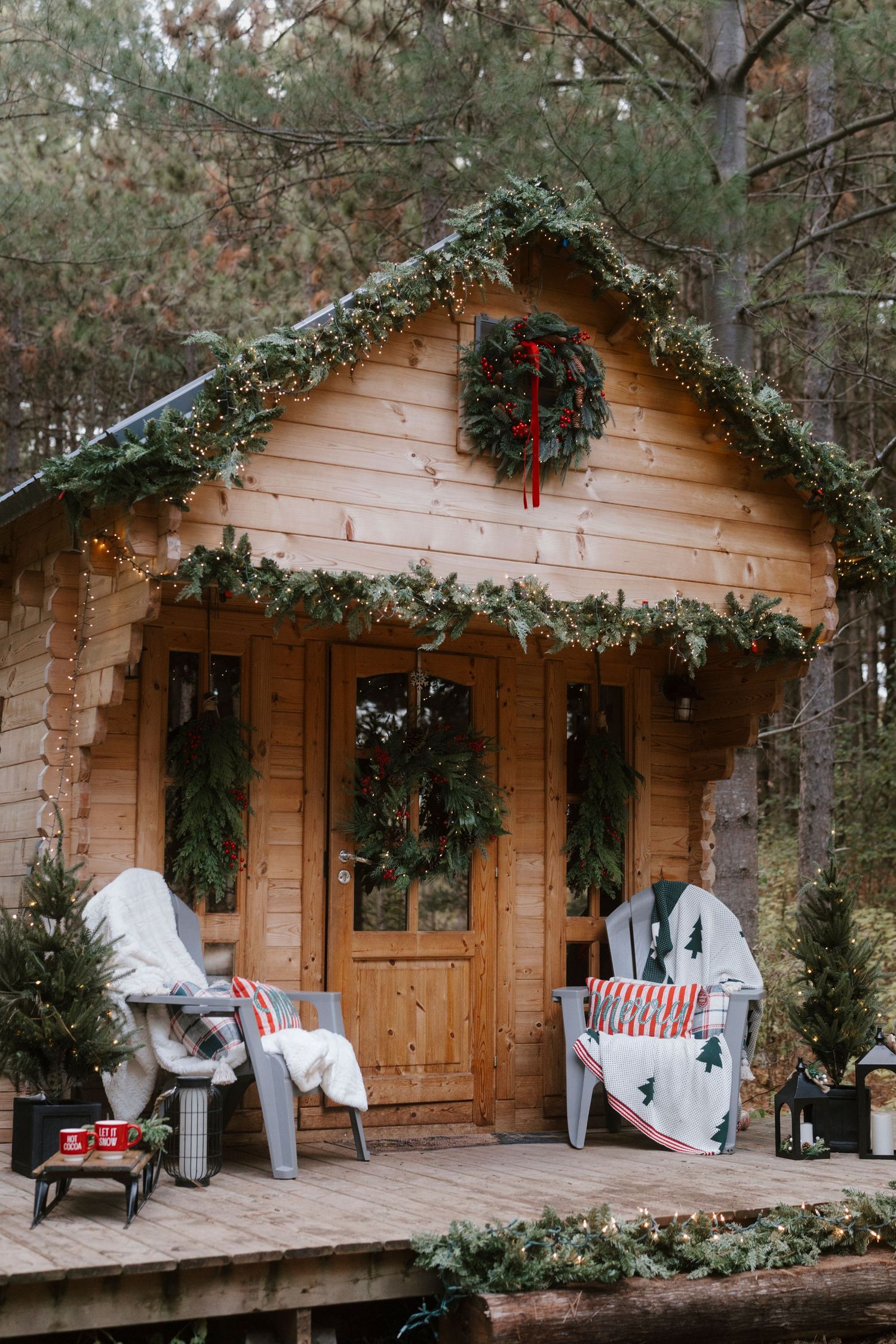 Cozy wooden cabin decorated with Christmas wreaths, garland, fairy lights, and Adirondack chairs on a festive porch in the woods.