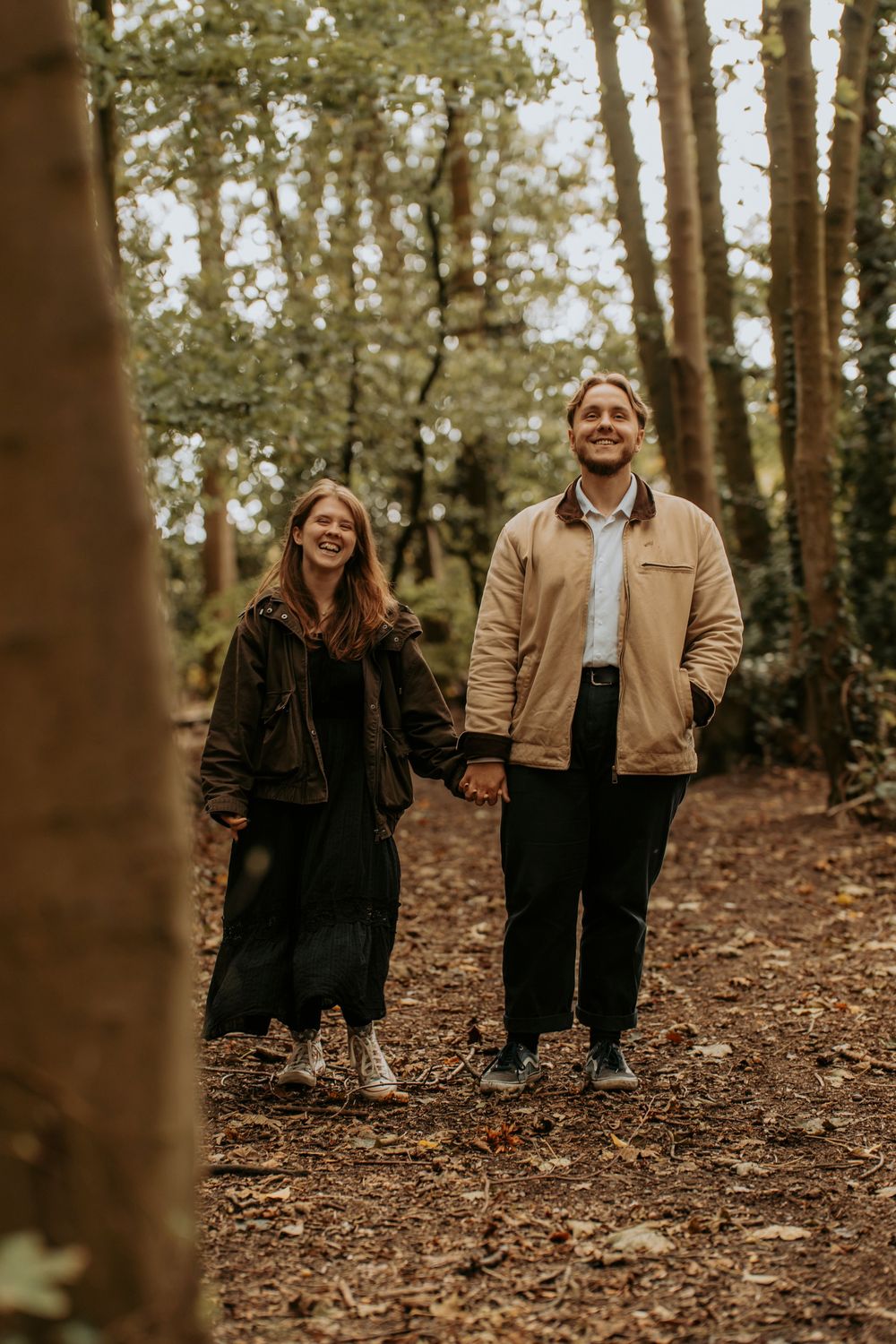 A smiling couple holds hands while walking on a leaf-covered forest path, wearing casual autumn jackets.