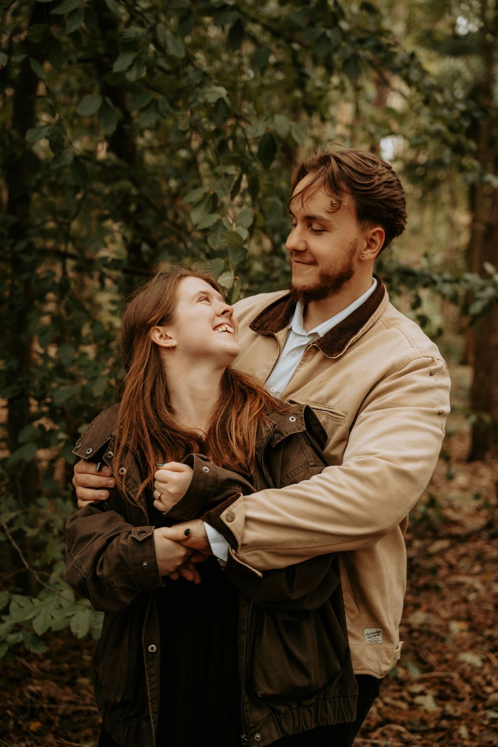 A smiling couple embracing in an autumn forest, woman laughing up at bearded man in tan jacket.