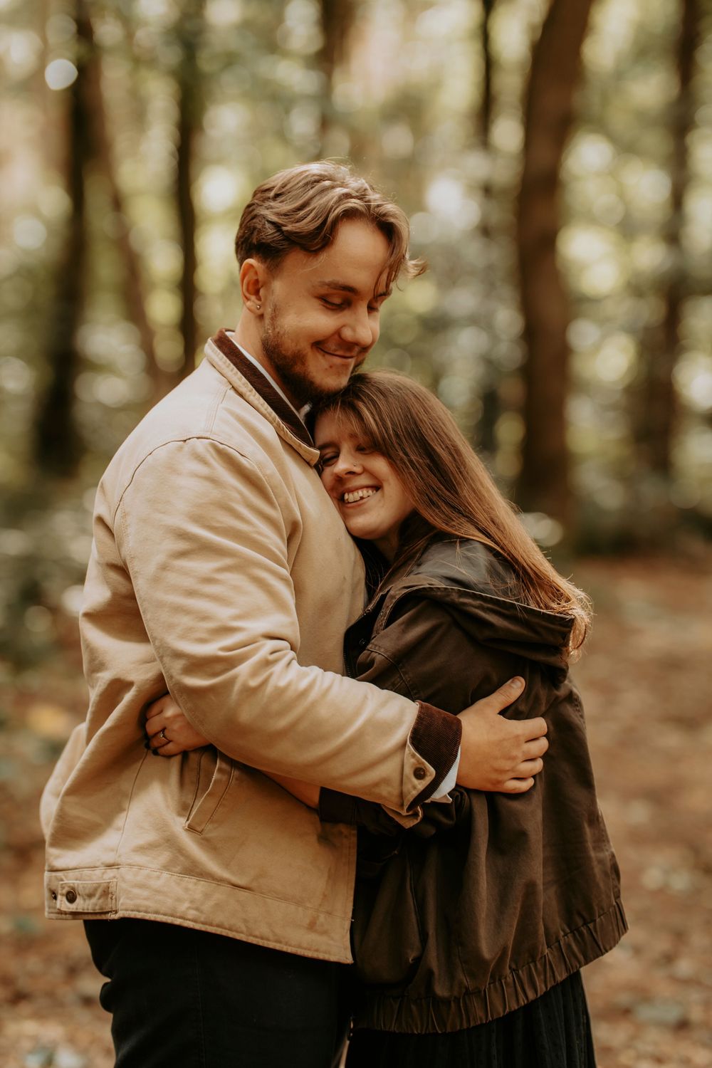 A couple embraces warmly in an autumn forest, smiling joyfully while wearing casual jackets among golden trees.