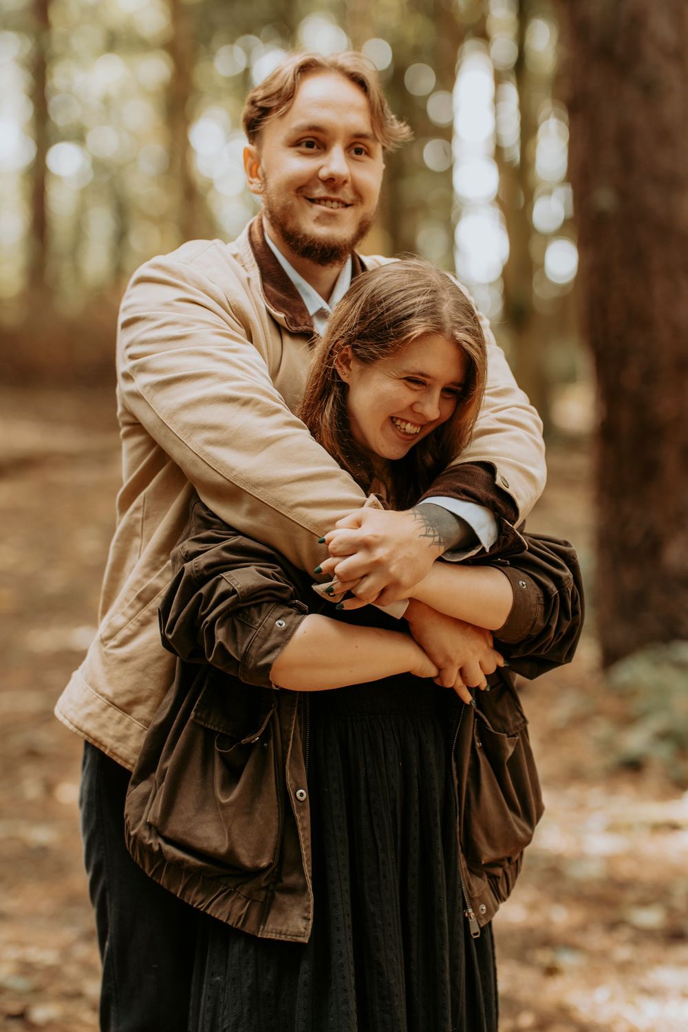 A smiling couple embraces in a forest, man in beige jacket hugging woman in dark coat among trees.