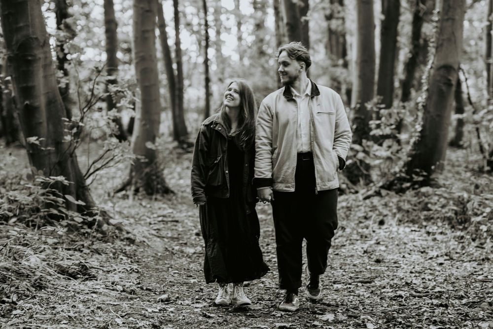 Couple holding hands on a forest path, smiling in black and white photo among tall trees.