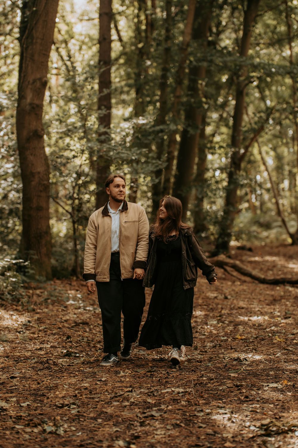Couple holding hands walking through a forest path covered in autumn leaves, surrounded by tall trees.