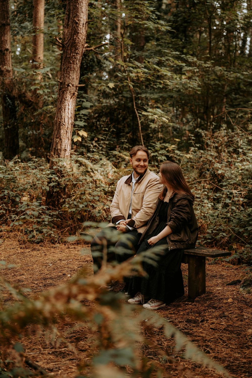 Couple sitting together on a wooden bench surrounded by lush green forest trees and autumn foliage.