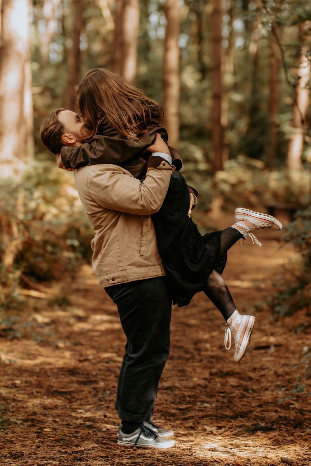 Couple sharing a romantic embrace in a sunlit forest, man lifting woman off the ground among tall pine trees.