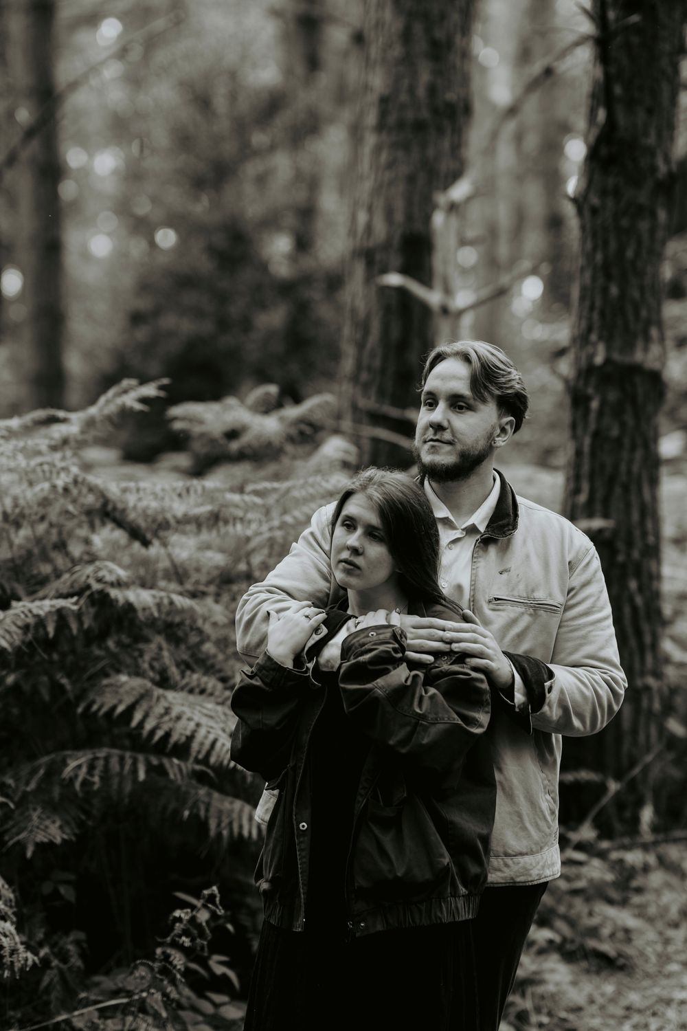 Black and white photo of a couple embracing in a lush forest, man holding woman from behind among ferns.