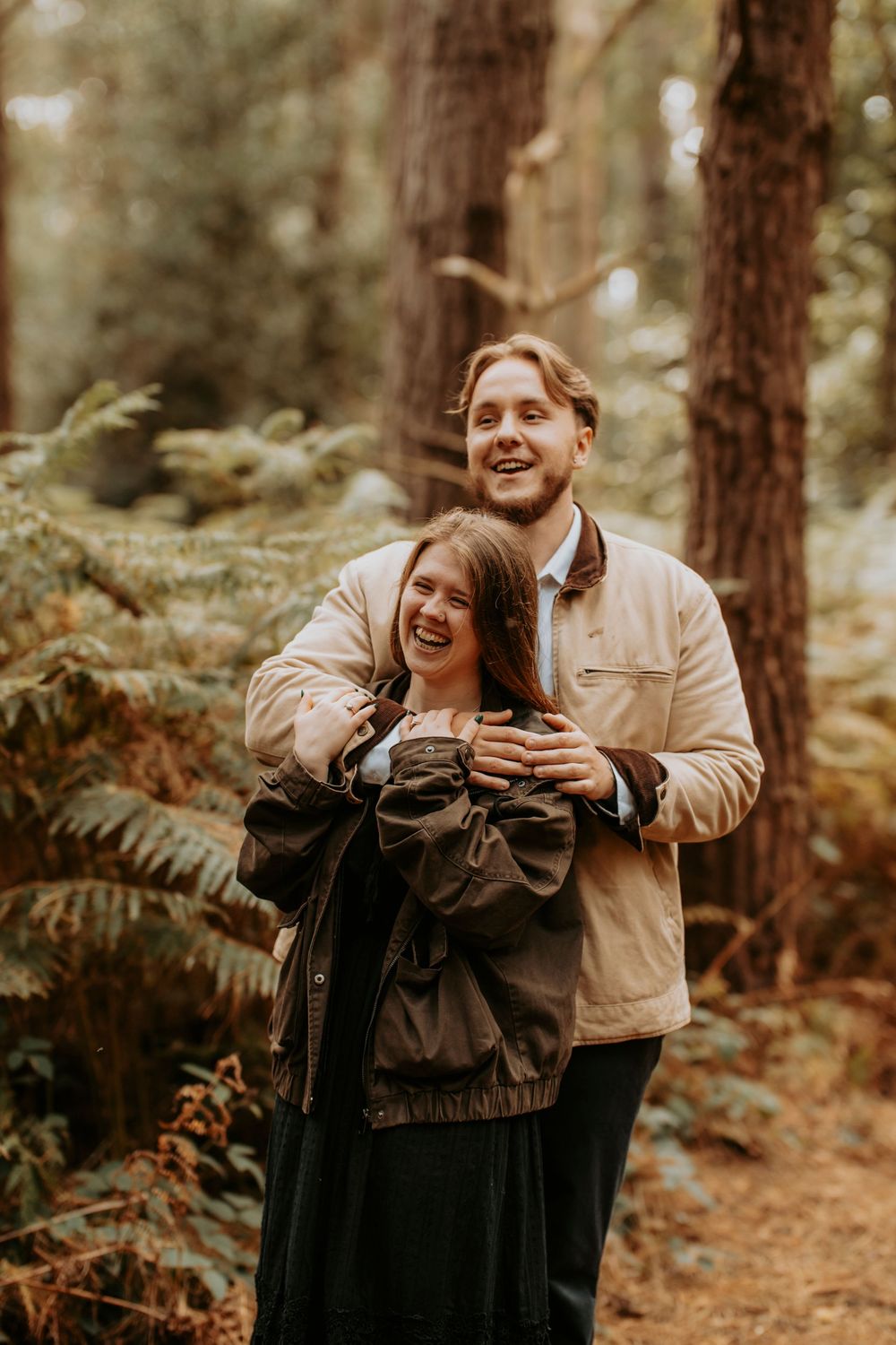 A laughing couple embraces in a lush forest, wearing casual jackets among tall trees and ferns.