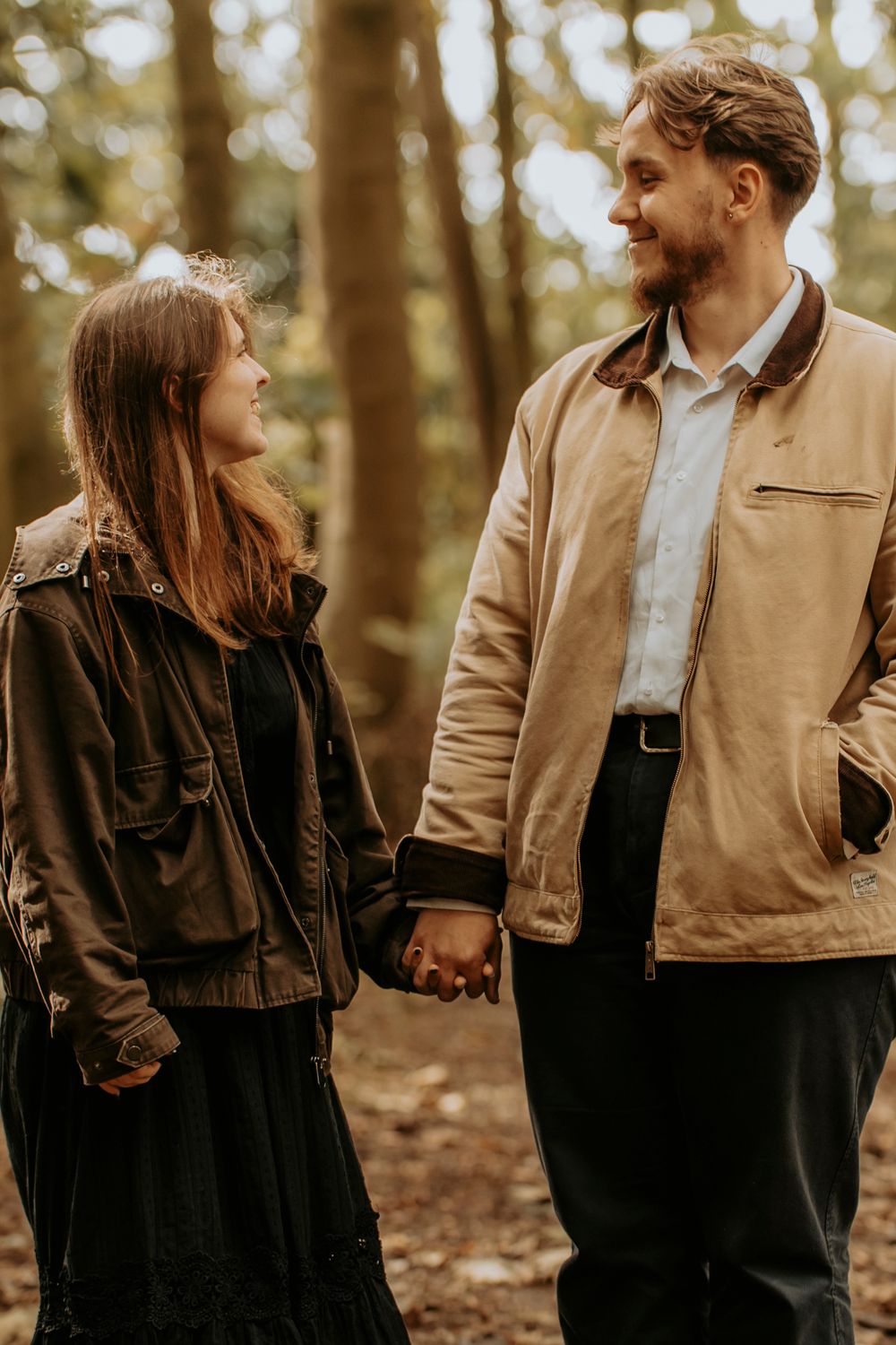 A couple holds hands and smiles at each other in a forest, wearing casual jackets in warm autumn tones.
