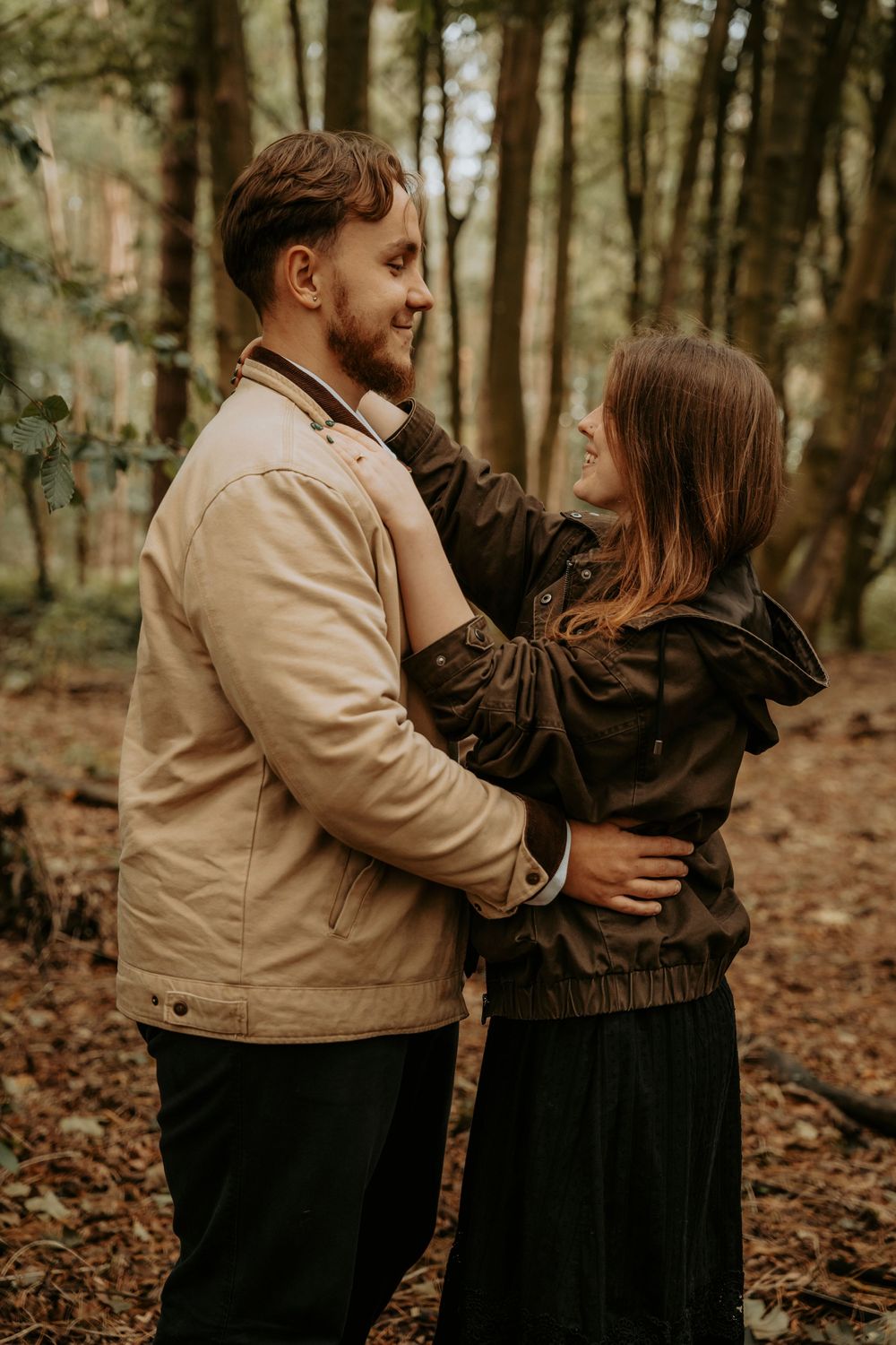 A couple embracing in an autumn forest, the man in a beige jacket smiling as the woman in black looks up at him.