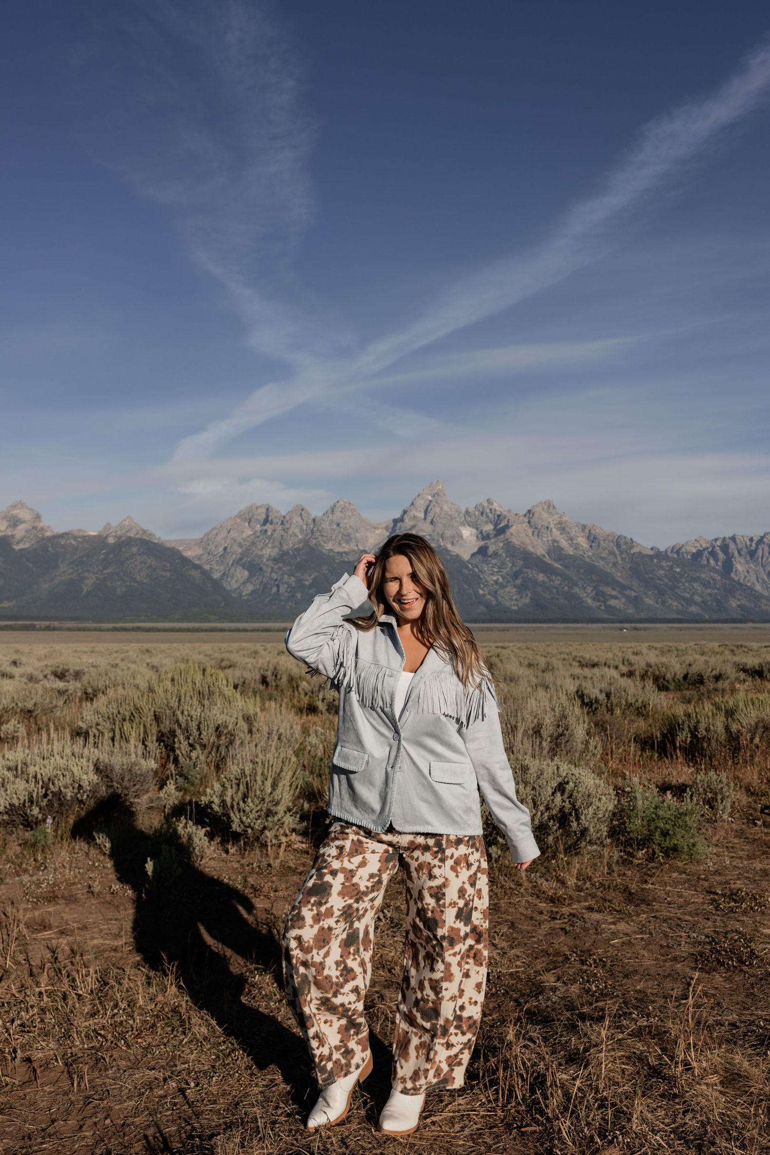 Woman in a light blue button-up top and cow print pants stands on a gravel path beside golden aspen trees in autumn.